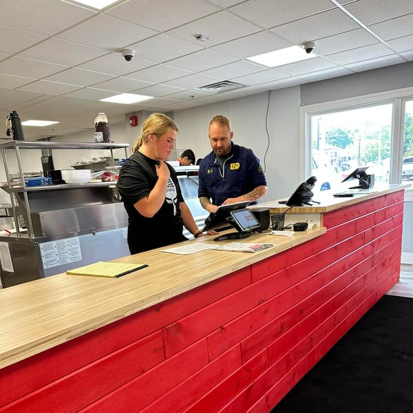 Two Home Slice Pizzeria employees, a man and a woman, stand behind a red and wood counter.