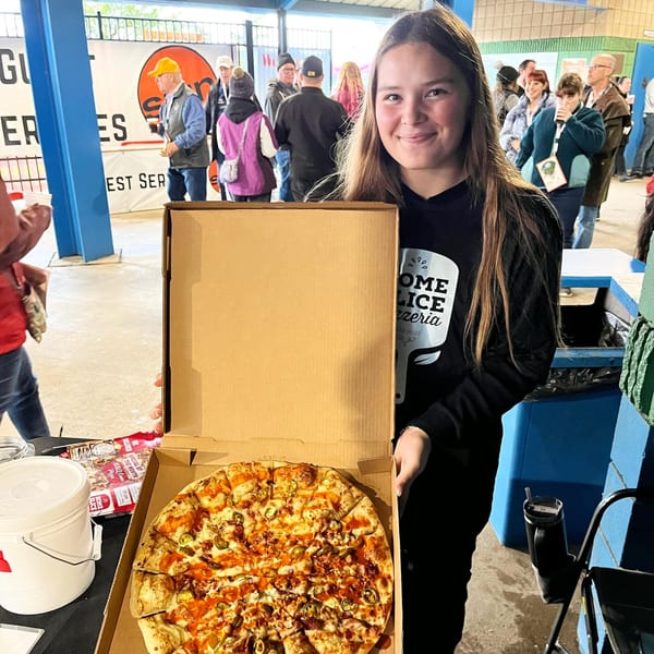 A smiling woman in a Home Slice Pizzeria shirt holds an open pizza box with a BBQ chicken pizza.