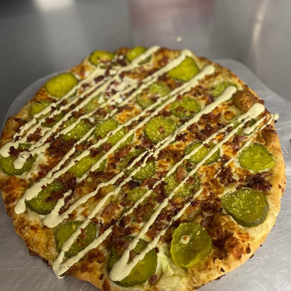 A Home Slice Pizzeria pizza topped with pickles, bacon, and a creamy white sauce drizzle.