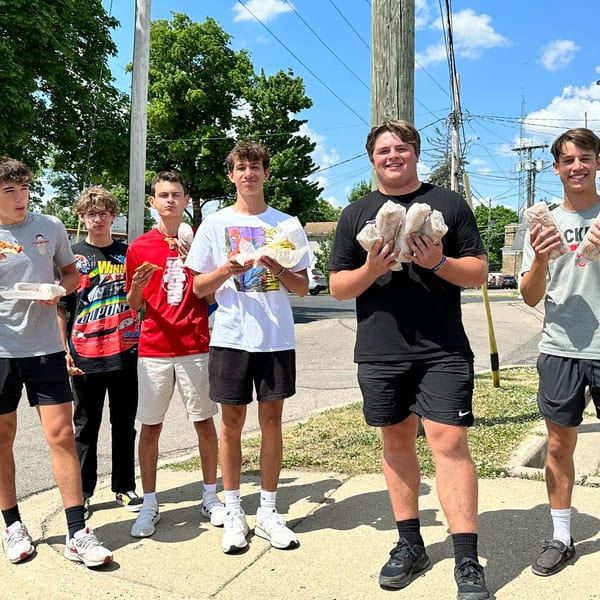 Six young men stand outside on a sunny day, holding pizzas and sandwiches from Home Slice Pizzeria.