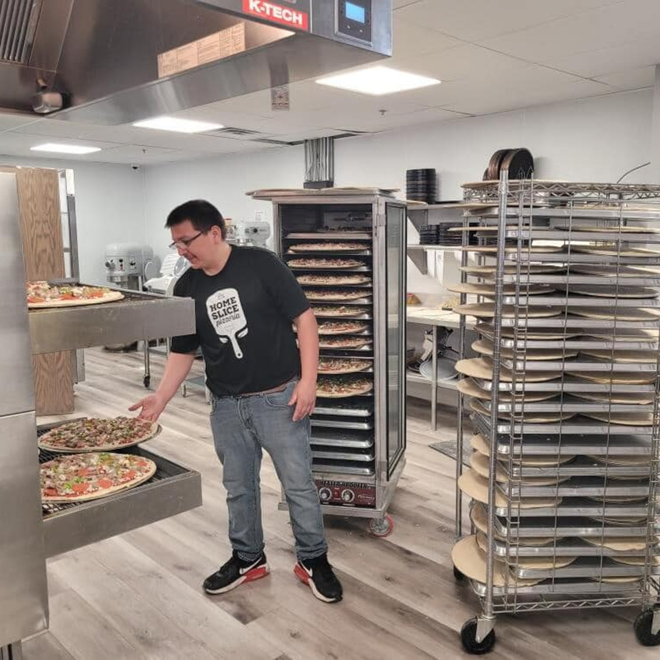 A Home Slice Pizzeria employee places an uncooked pizza into a large commercial oven.